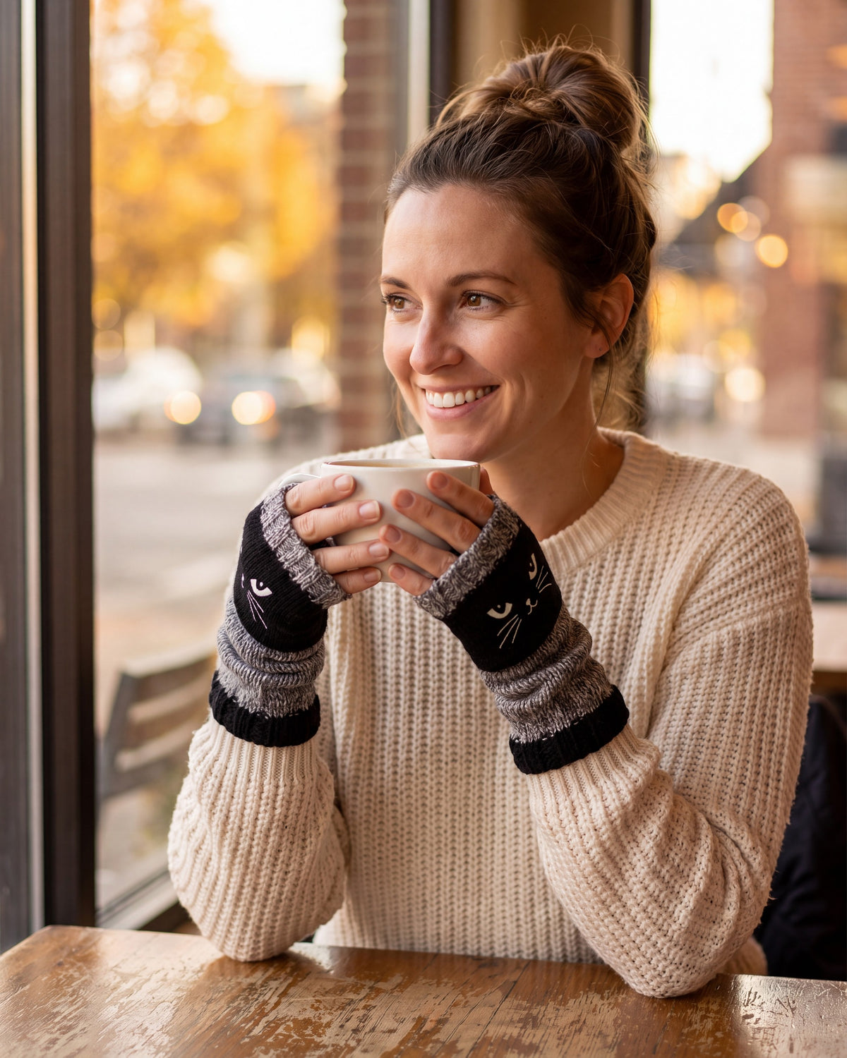 Woman in a cafe holding a latte while wearing black fingerless gloves with an embroidered cat face