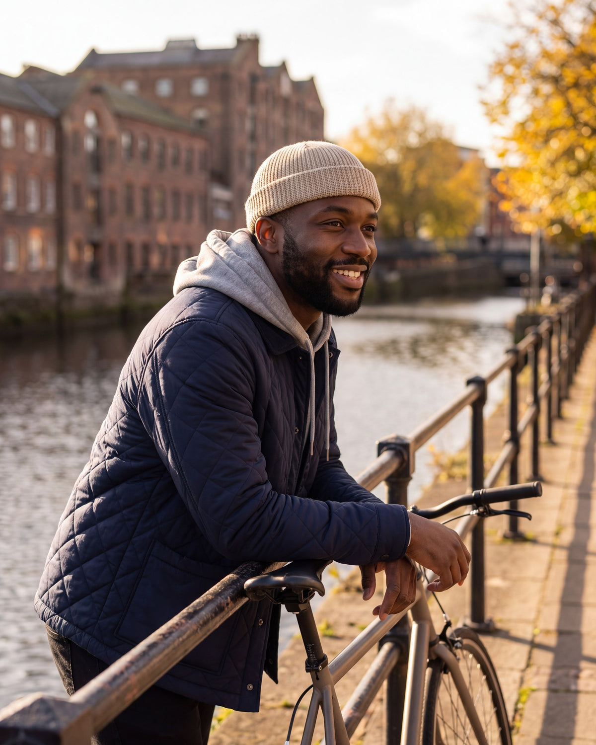 Man in a khaki fisherman beanie leaning on a riverside railing next to his bike