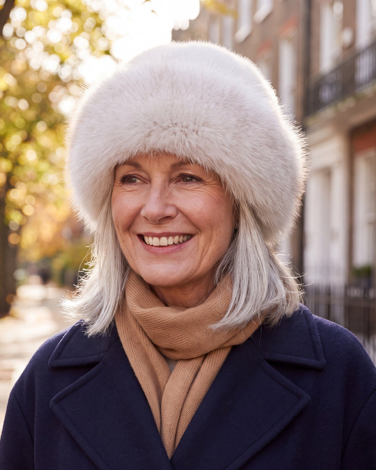 Smiling woman with silver hair wearing a milky white faux fur hat with rolled brim on a city street