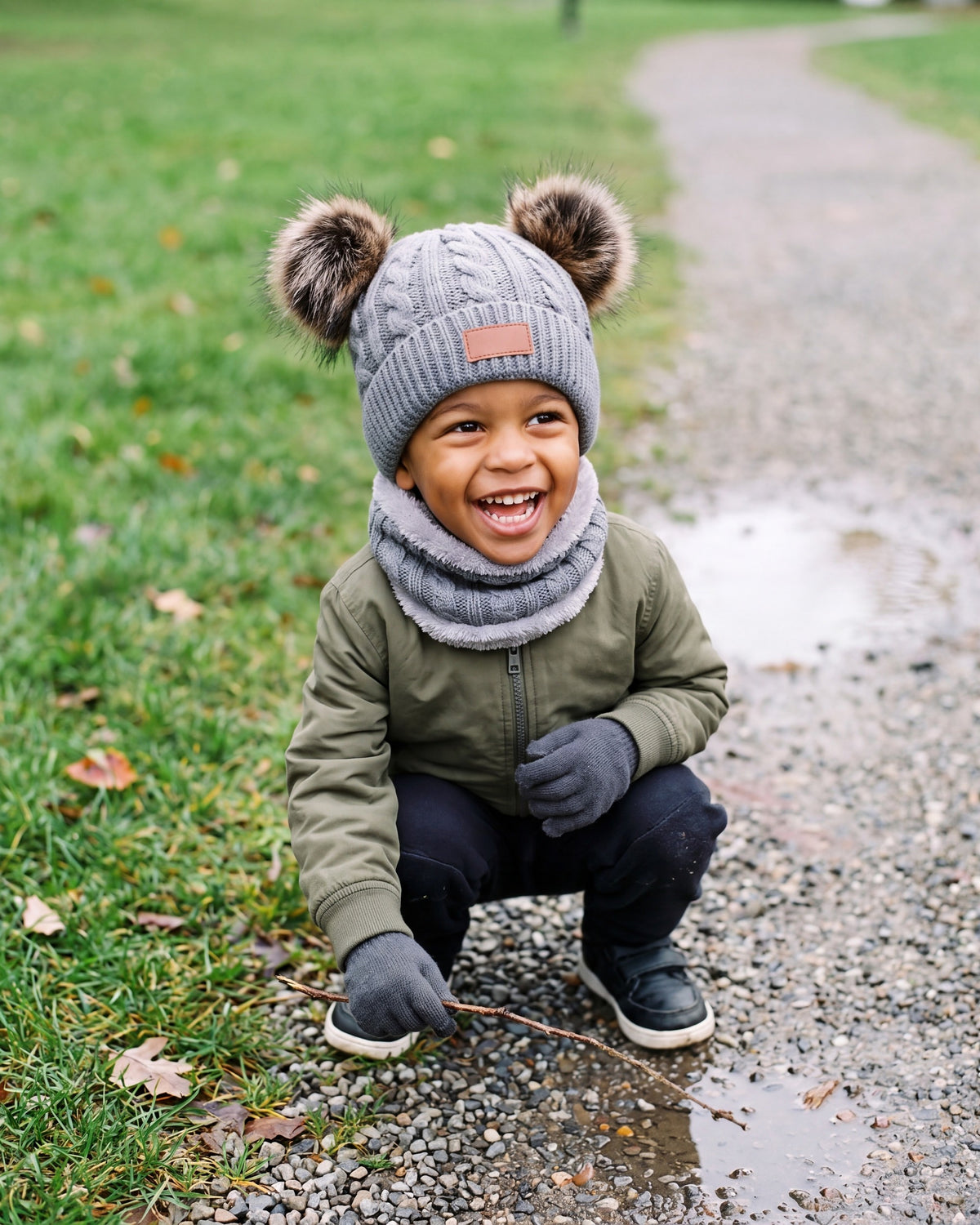 Laughing boy crouching on a park path wearing the gray pom pom beanie, neck warmer and gloves set