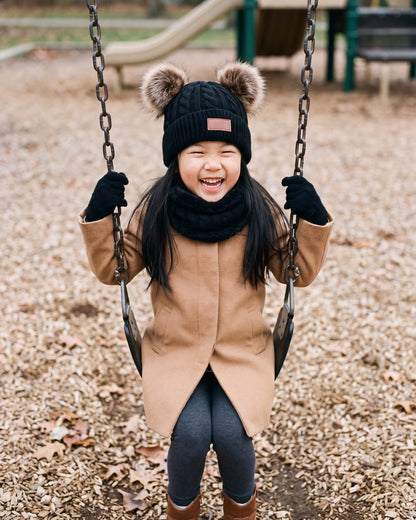 Girl laughing on a playground swing wearing the black pom pom beanie, neck warmer and gloves set