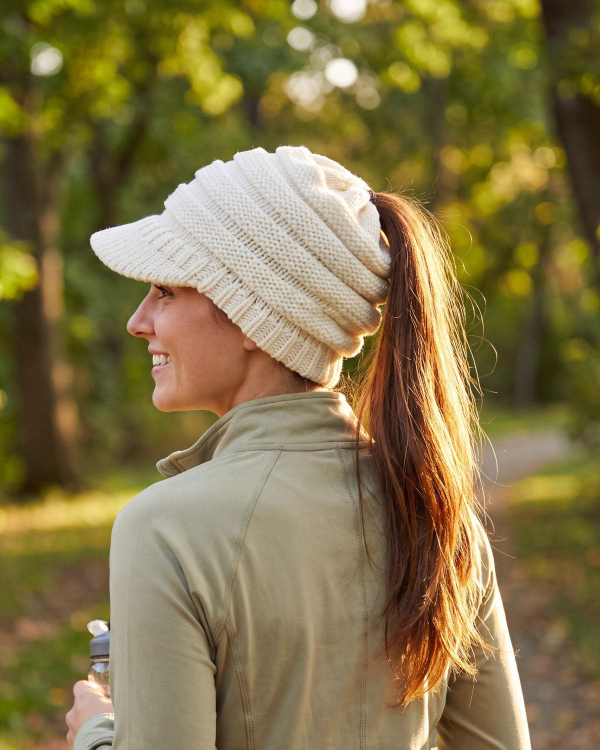 Woman walking in a park wearing a white ponytail beanie with brim, ponytail through the back